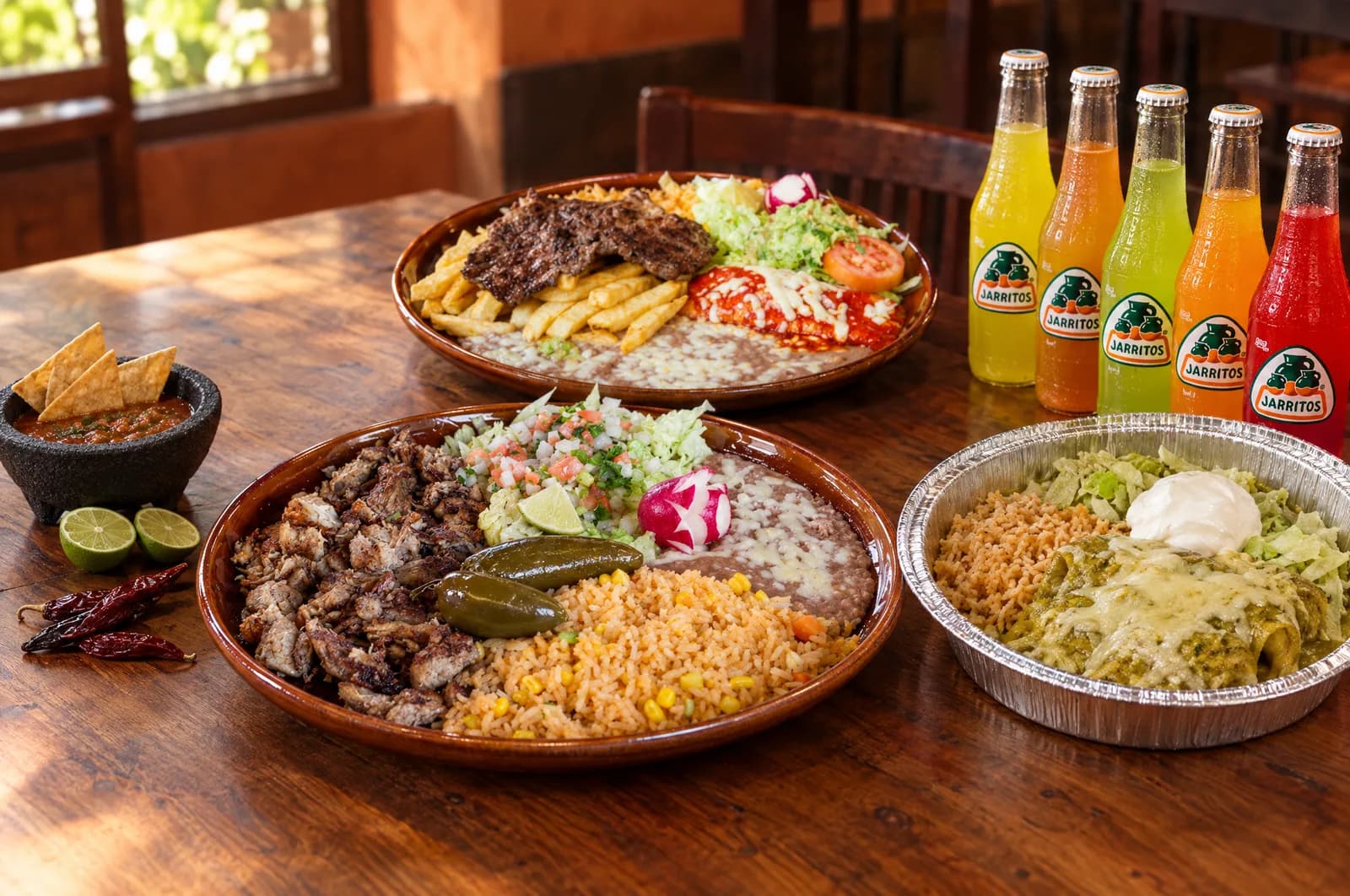 Table spread with a carnitas plate, a tampiqueña plate, a tray of enchiladas verdes and five Jarritos sodas, with chips and salsa
